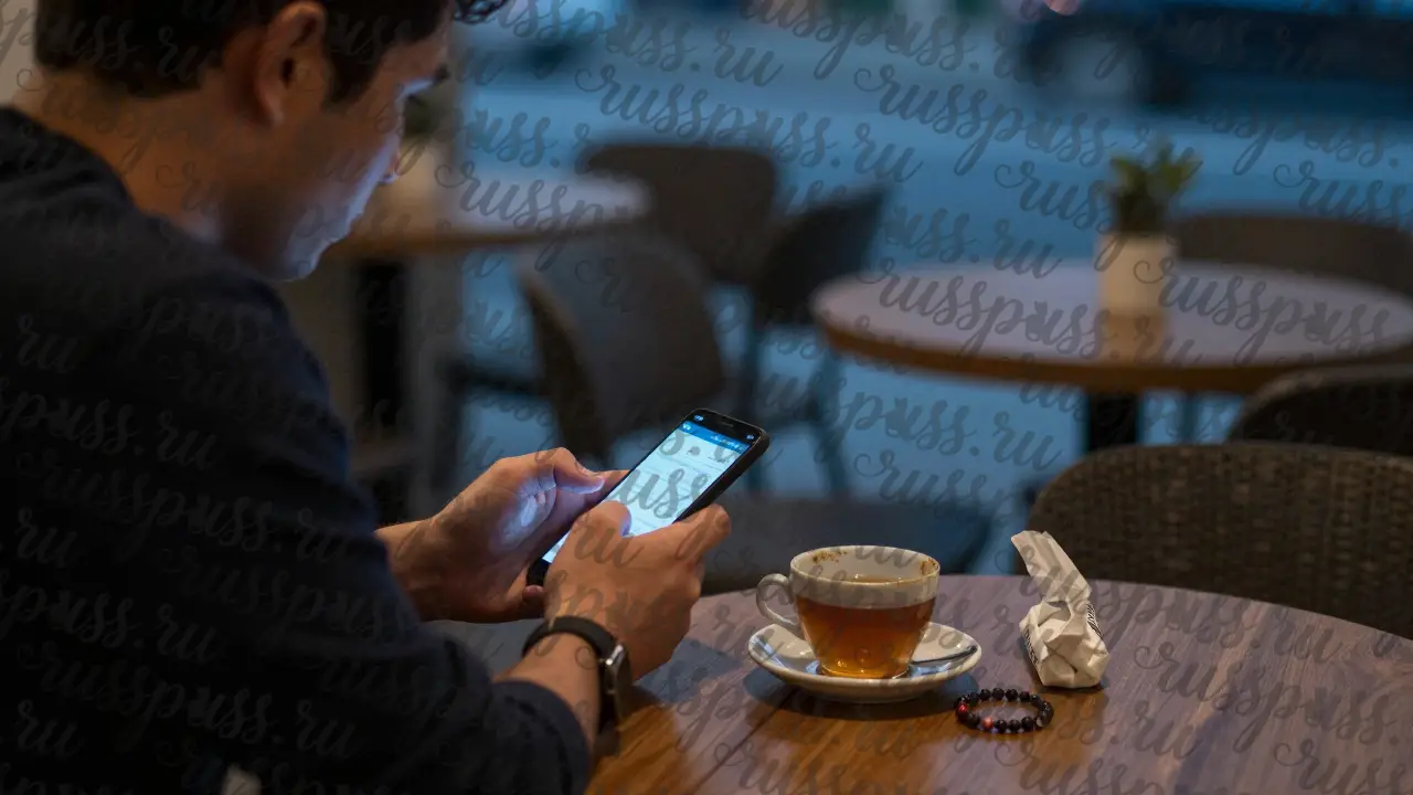 Мужчина смотрит на телефон с поздним сообщением, рядом — непитый чай и браслет, тусклый свет ночи.